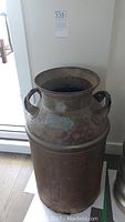 Full view of vintage metal milk can showing cylindrical body, wide rim, and dual handles, placed indoors on wooden floor.