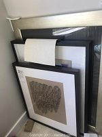 Two black framed artworks leaning upright; one partially visible feather pen drawing signed by W.M. Smith Jr., another colorful shadow box framed artwork behind.