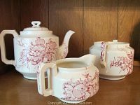 Photo showing ceramic teapot, sugar bowl, and creamer with red botanical design against wooden background
