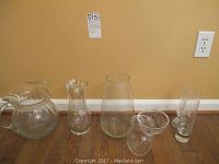 Six clear glass items arranged on floor against wall including pitchers and vases in various shapes and sizes.