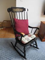 Black wooden rocking chair with gold floral decoration on backrest, ribbed red back cushion and floral seat cushion, showing wear on wood.