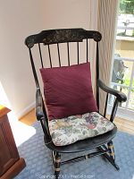 Front angled view of the black wooden rocking chair with burgundy back cushion and floral seat cushion under natural light.