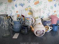 Full view of ceramic pitchers, teapots, cream and sugar containers in various colors and patterns on countertop.