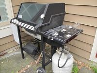 Photo of the full Weber gas grill positioned outdoors against a beige siding wall, showing the grill body, control panel with three knobs, and attached propane tank below.