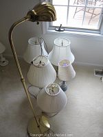 Tall bronze painted standing lamp with curved neck and dome-shaped shade next to five table lamps with white shades of varying shapes and bases on a beige carpet near window lighting.