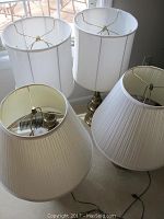 Two pairs of table lamps near a window, one pair with white drum-shaped shades and brass bases, the other with pleated ivory shades and dark bases.