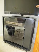 Media cabinet TV stand with glass front doors shown closed with electronic equipment visible inside through the glass and a large flat screen TV placed on top.