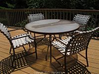 Outdoor glass top round patio table with rusted metal frame surrounded by four white woven strap patio chairs on wood deck in sunlight.