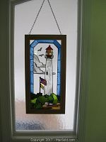 Front view of framed stained glass art showing Cape May lighthouse with blue border, clouds, birds, flags, and shrubbery.