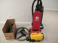 Red Dirt Devil upright vacuum with hoses and other vacuum accessories, including a yellow Eureka Mighty Mite compact canister vacuum and a green Hoover upright vacuum in background.