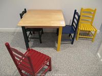 Photo showing entire children's table with four different colored wooden chairs around it, displayed on carpeted floor.