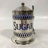 Front view of silver plated sugar container with hammered texture, blue glass inlays, and 'SUGAR' cut-out lettering.