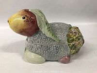 Side view of ceramic rabbit container showing textured gray body, yellow head with brown shading, and green leaf details on ears and feet.