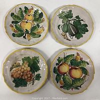 Four round faience pottery plates with different hand-painted fruit motifs arranged in a square, white background, yellow rim with scalloped edges.