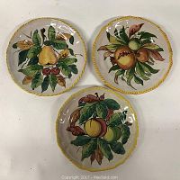 Three Italian faience pottery plates displayed flat showing various fruit motifs and yellow twisted edge decoration.