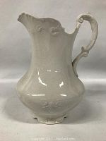 White ceramic ironstone pitcher with ornate handle and scalloped spout against plain background.