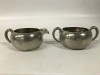 Front view of both pewter containers: creamer with spout on left and sugar bowl with two handles on right, both with 'D' monogram.
