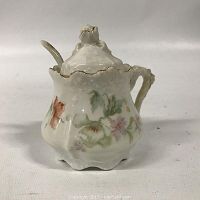 Front view of antique German mustard pot showing floral decorations and scalloped edges with lid on.