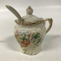 Front view of closed antique mustard pot showing floral decoration and gilt edges with spoon inserted