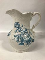Side view of large white ceramic pitcher with ornate handle, scalloped rim, blue floral transferware decoration, and embossed details.