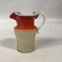 Front angle showing amberina glass pitcher with textured lower half and smooth red-orange upper half, cream handle, and flared lip.