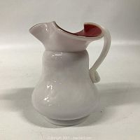 Front facing view of a small cased glass pitcher showing white outer glass with pink inner highlights at the rim and spout, curved handle, slightly flared base.