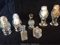 Group of antique glassware salt and pepper shakers, small tincture bottle, and square salt cellar on black fabric background.