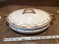 Oval covered serving dish with gold-tone handles and lid handle on a carpeted floor, measuring about 12 inches long.