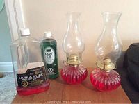 Photo showing two red glass oil lamps with brass burners and clear chimneys, plus two bottles of lamp oil on a wooden surface.