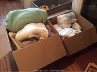 Two cardboard boxes filled with assorted linens including blankets, towels, tablecloths, and napkins as seen from above.