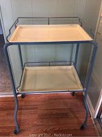 Full view of the gray metal frame medical instrument cart with two cream-colored removable enamel trays and four wheels on hardwood floor.