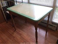 Full view of enamel kitchen table showing light-colored enamel top with green trim and turned wooden legs on a hardwood floor.