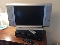 Silver 19 inch Venturer LCD TV with remote on top of black Sharp VHS player, sitting on wooden surface with lamp in background.