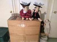 Two stuffed Christmas moose sitting on a large cardboard box with other decorations beside it