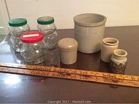Photo showing all items together on surface: four crockery containers of varying sizes, three glass peanut butter jars with colored lids, and two wooden yard sticks.