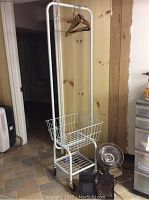 Metal laundry rack with wire basket and wheels, holding wooden hangers, alongside 12 inch table fan on tiled floor