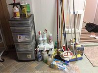 Wide shot showing the full collection of cleaning supplies and tools against a wall: multiple spray bottles, plastic storage container with drawers, mops, brooms, and cleaning cloths.