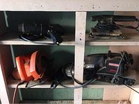 Shelf holding four power tools including BD circular saw, BD sander, Craftsman belt sander, and a paint stripping heater