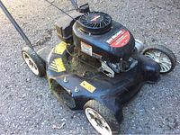 Front-side view of black Yard Machines push lawn mower with engine and wheels visible, showing grass residue and missing grass discharge cover.