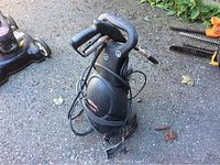 Black Jobmate electric pressure washer with power cord and hose coiled around it, standing on asphalt outdoors near other lawn tools.