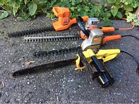 Full view of four power gardening tools: two Black & Decker electric hedge trimmers, one Sunbeam electric hedge trimmer, and one electric chain saw, all placed on an outdoor surface.