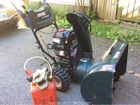 Front right view of green Craftsman two-stage gas snow blower with manual chute and knobby wheels. Includes plastic gas can and gloves.