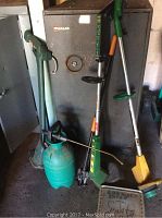 Photo showing three electric weed whackers, green spray can with brass wand, and a seed spreader with green handle.