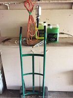 Full view of green dolly with two containers of washer de-icer fluid and a handheld trouble light with orange extension cord on a white counter.