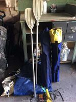 Photo of uninflated blue and white dinghy with two aluminum paddles and two portable lawn chairs in blue and yellow carrying bags inside a garage or storage room.