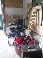 Photo showing two walkers, a blue wheelchair, wooden crutches, red plastic bin with metal parts, and a black carrying case.