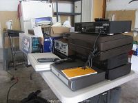 Table with stacked electronic items including black DVR boxes, a remote, and a printer in background; shows overall lot contents