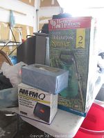 Boxed multipurpose garden sprayer next to AM/FM speaker box and tissue box in garage setting.