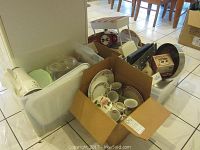 Boxes filled with assorted kitchenware including plates, mugs, cooking pans and trays.