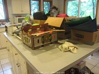 Wide view of kitchen countertop showing boxes with assorted kitchen items including pots, pans, textiles, serving platters, and other items.